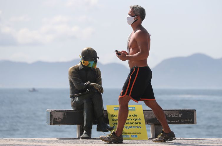 Um homem caminha ao lado da estátua do escritor brasileiro Carlos Drummond de Andrade que usa máscara protetora no primeiro dia de uso obrigatório de máscaras  em meio ao surto de doença por coronavírus (COVID-19), na praia de Copacabana